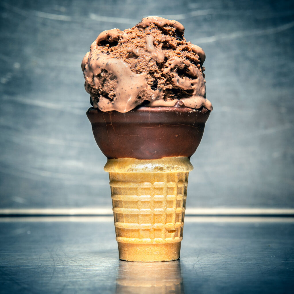 An ice cream cone containing a scoop of chocolate ice cream with fudge pieces and a fudge swirl.