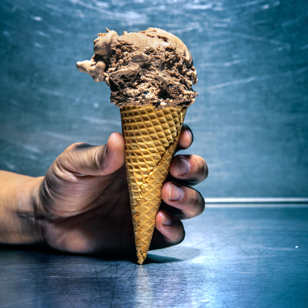A hand holding an ice cream cone containing a scoop of chocolate ice cream with cookie pieces and a fudge swirl.