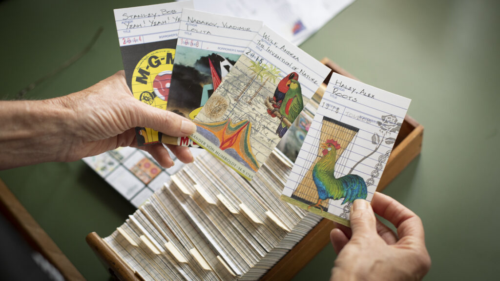 Hands holding art cards depicting four books including Roots, Lolita, and The Invention of Nature.