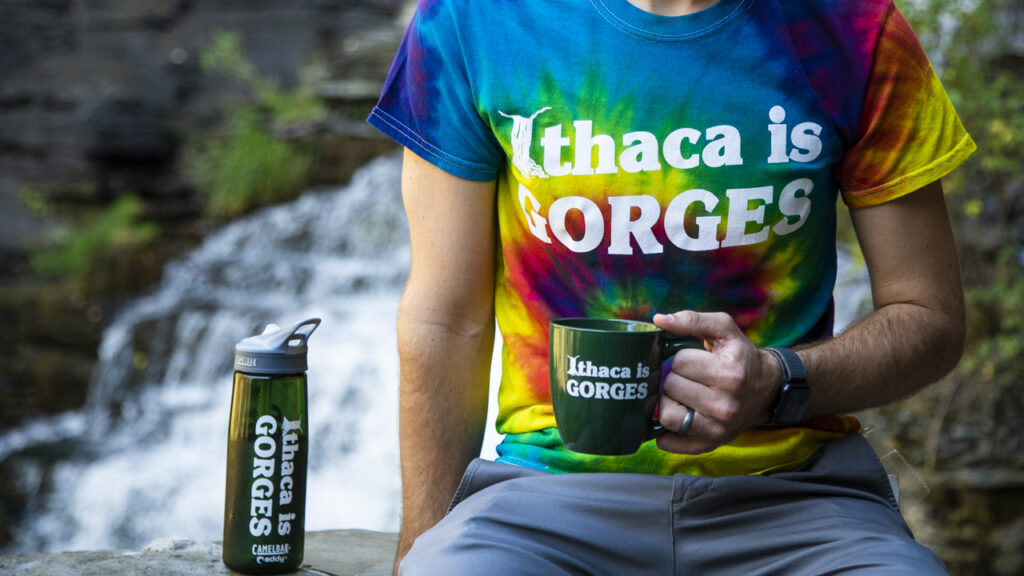 A man in a tie-dye T-shirt, holding a coffee cup, sitting next to a water bottle (all with the slogan "Ithaca is Gorges") in front of. waterfall.