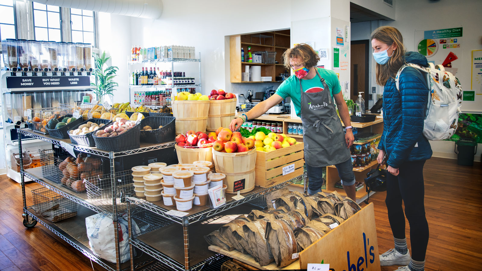The interior of a small grocery shop with a worker in an apron and a customer wearing a backpack, looking at produce.