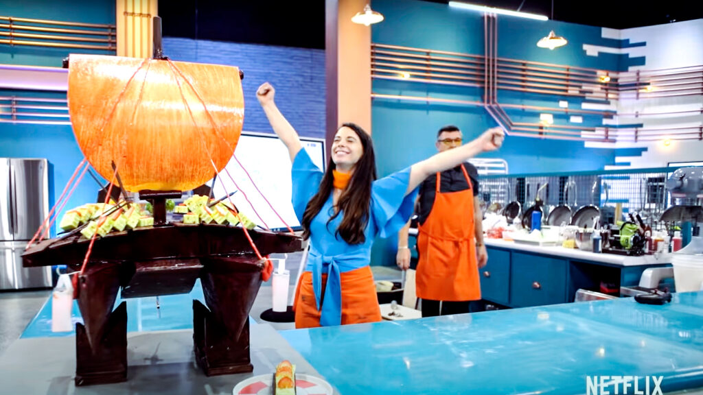 Renee Frohnert, MEng ’19, is shown in a scene from the trailer for the new Netflix show "Baking Impossible." At left is the double-hulled, chocolate-tempered, cake-based boat that she and her teammate, Steve, created for the first challenge
