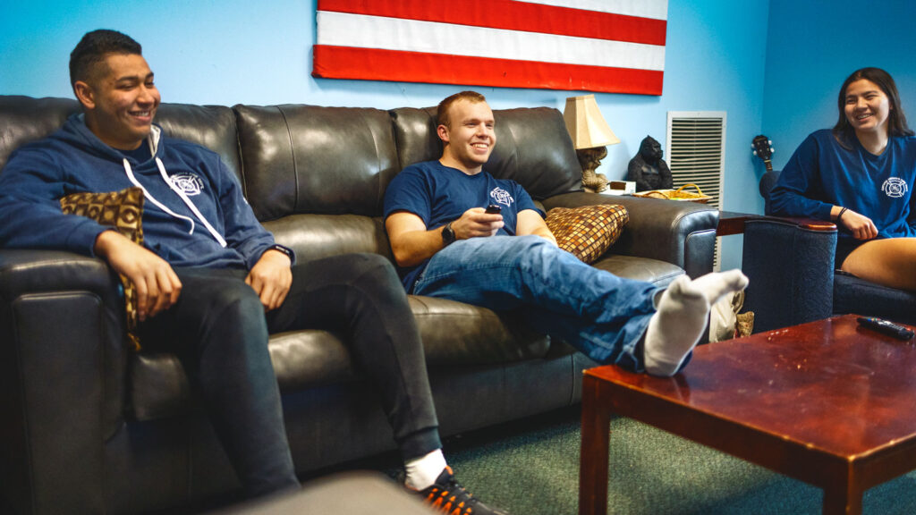 Three people sitting in a lounge. The man in the center has his feet up on the coffee table.