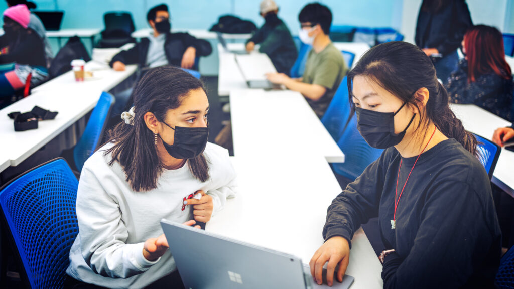 Two female students in masks looking at a laptop in a room with other students doing the same