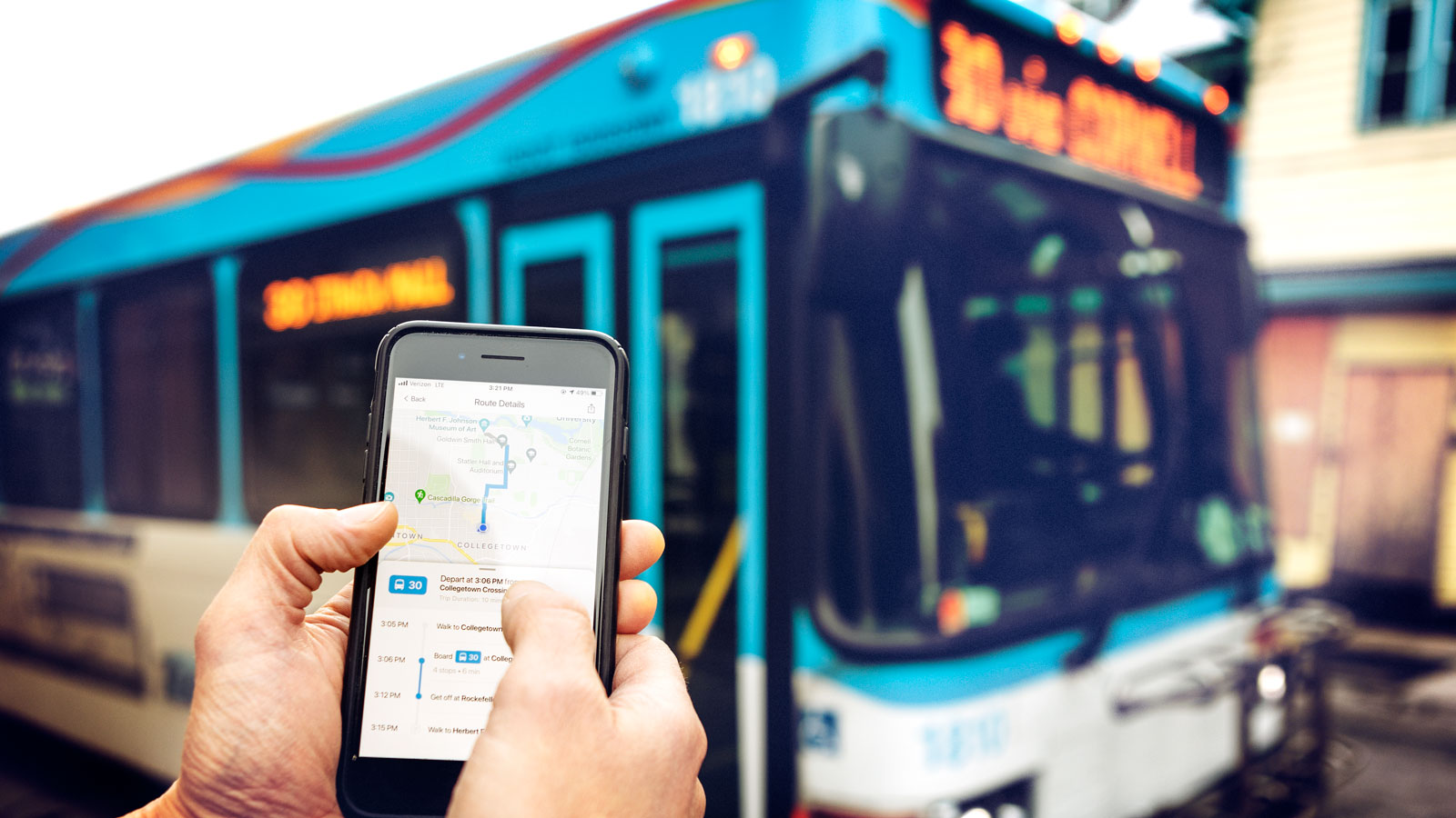 Hands holding a smartphone with the Ithaca Transit app on the screen with a bus behind it