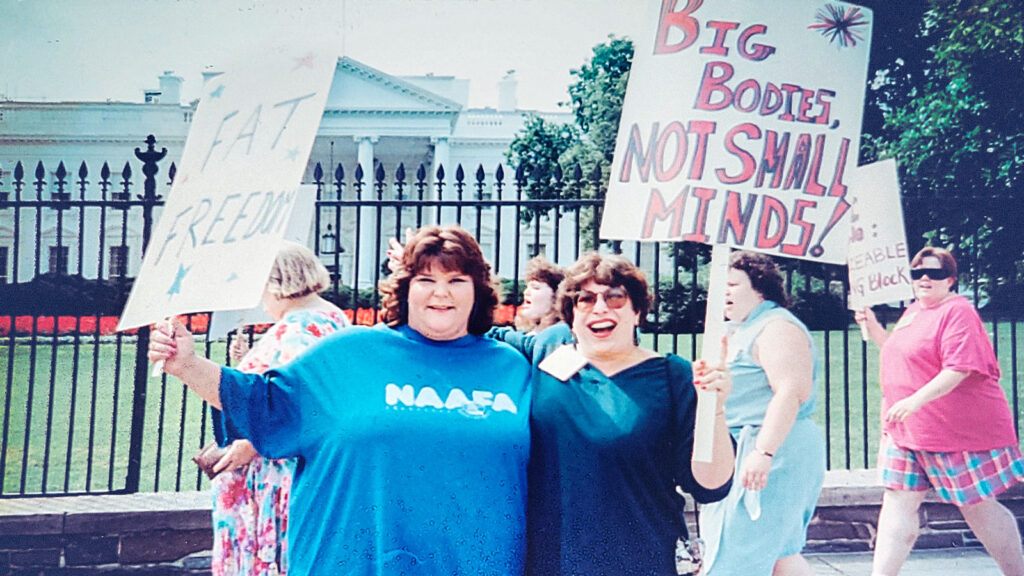 Two women holding protest signs in favor of size acceptance, in front of the White House