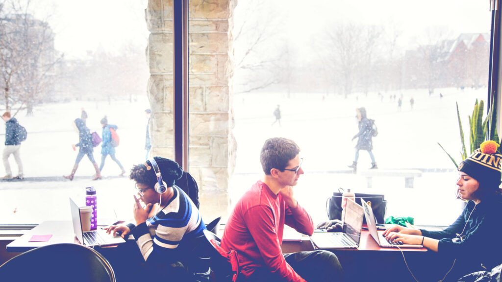 Students study in Olin Library as students walk across the Arts Quad in the snow outside