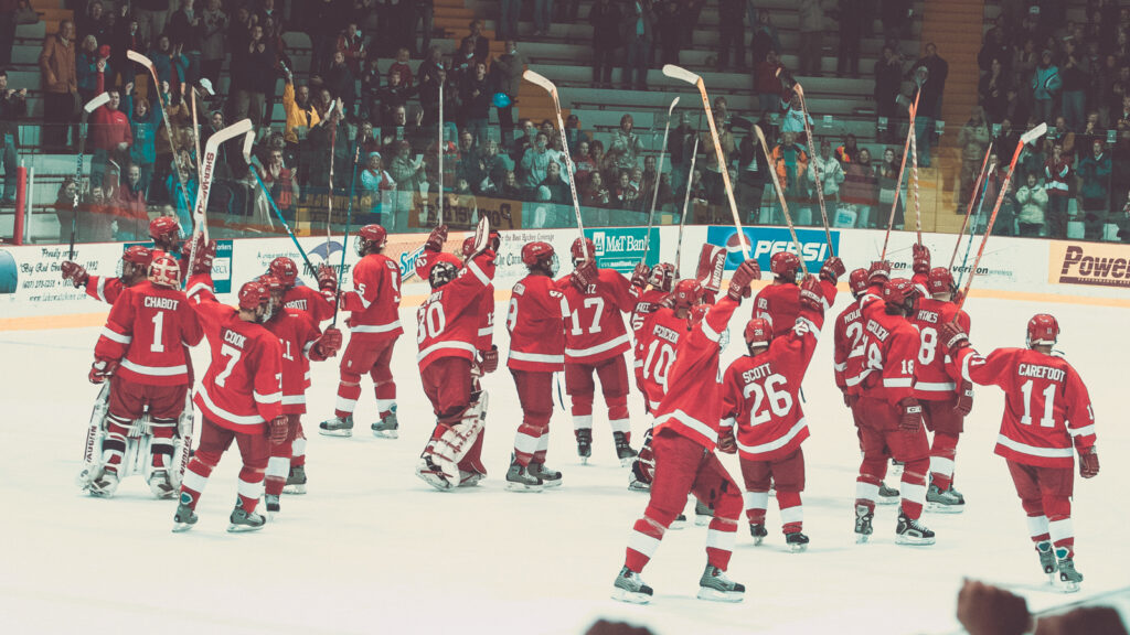 Cornell men's hockey players dressed in red jerseys raise their sticks at the end of the game to honor the crowd.