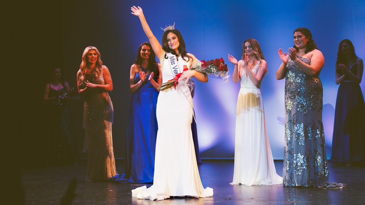 Alexina Federhen winning the Miss Vermont competition wearing a white dress and a tiara, holding a bouquet of flowers, with other contestants applauding behind her.