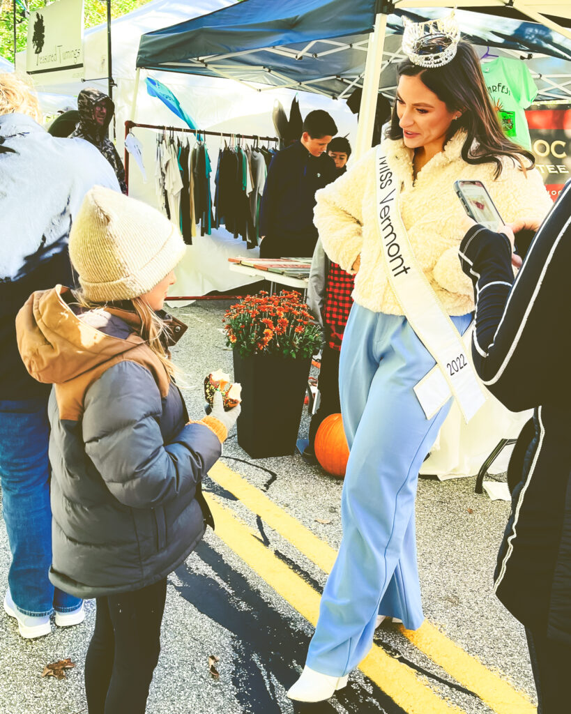 Miss Vermont wearing a tiara and white sash has a conversation with a child wearing a winter hat and coat at an event.