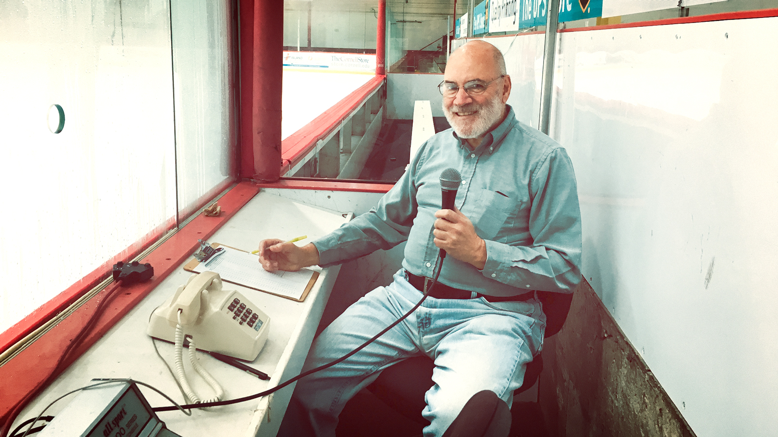 Cornell men's hockey announcer Arthur Mintz in the announcer's booth with a microphone.