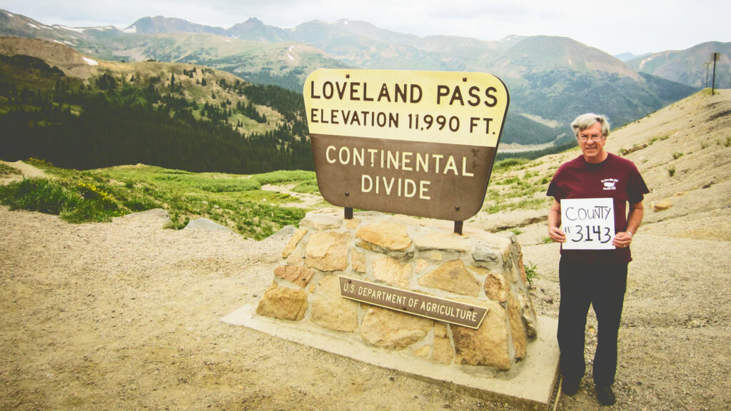 Brian O'Connor holding a sign that says "County #3143" next to a sign marking the Continental Divide at Loveland Pass, elevation 11,990 feet.