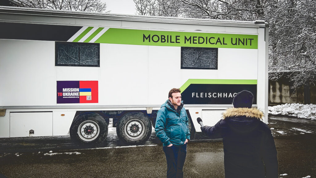 A man being interviewed by a TV journalist in front of a mobile medical van.