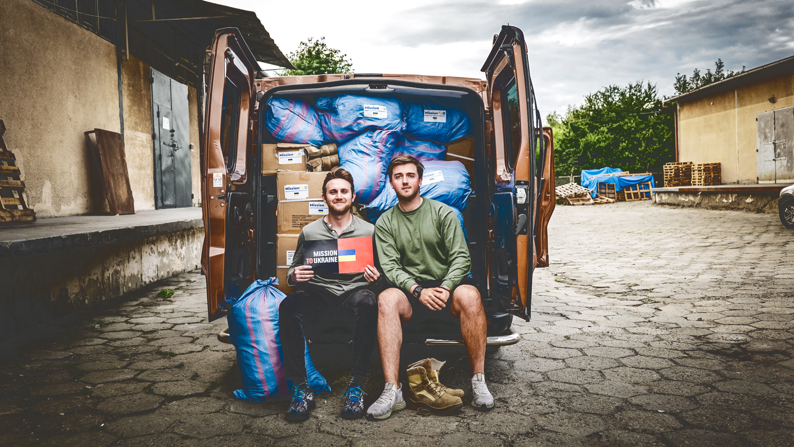 Two men sit on the back bumper of a van filled with packages