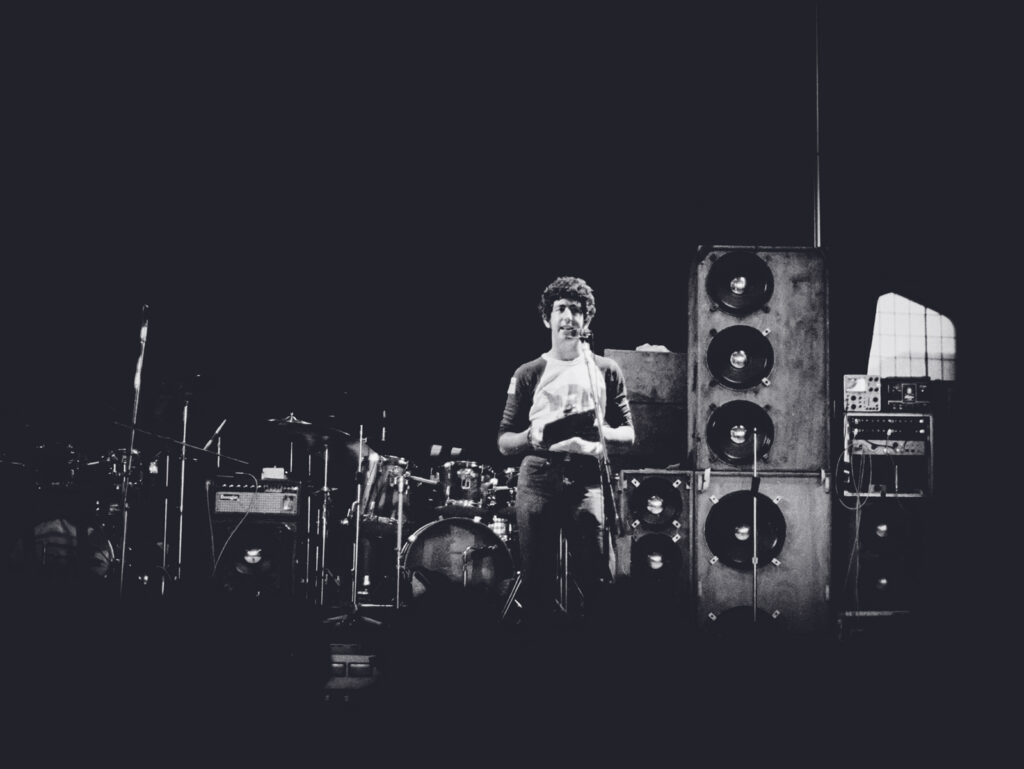 A college student stands on a stage with amplifiers next to him and a drumset behind him.