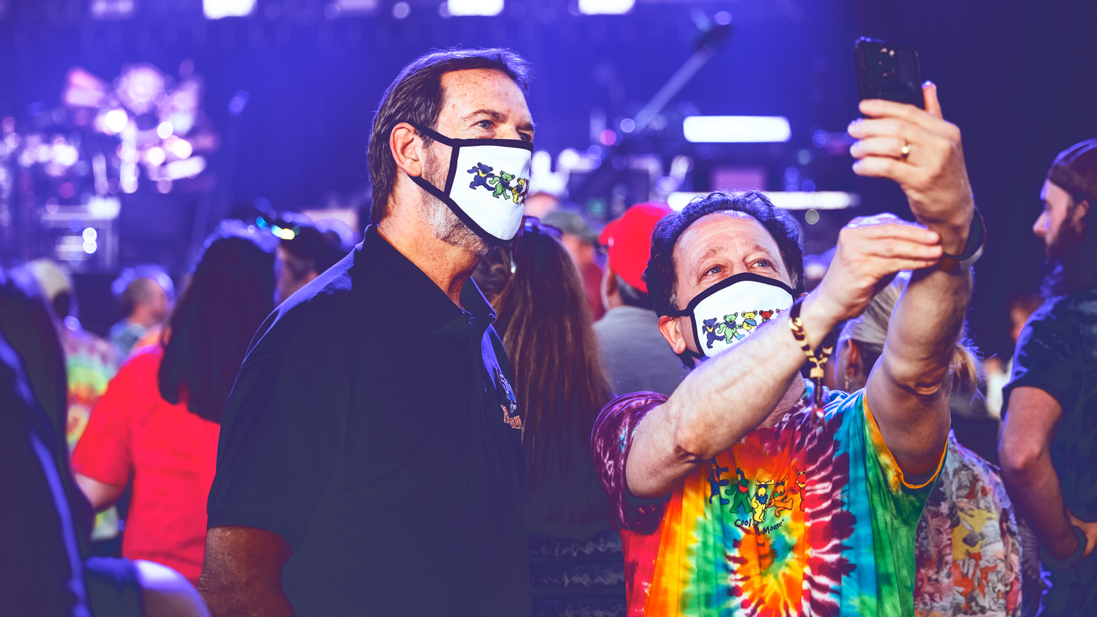 Two men wearing face masks with colorful dancing bears on them pose for a selfie at a concert.