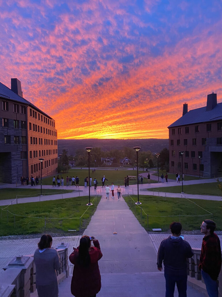 Four students gather to view and take pictures of an orange, yellow, and pink sunset at Cornell University.