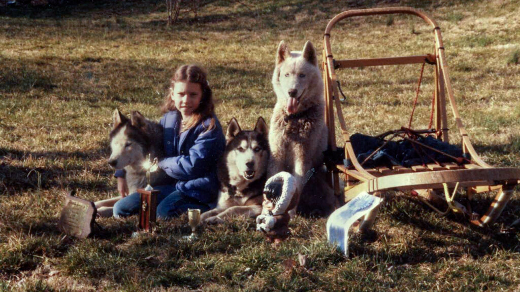 Heather Huson as a child with three sled dogs, a sled, and a blue ribbon