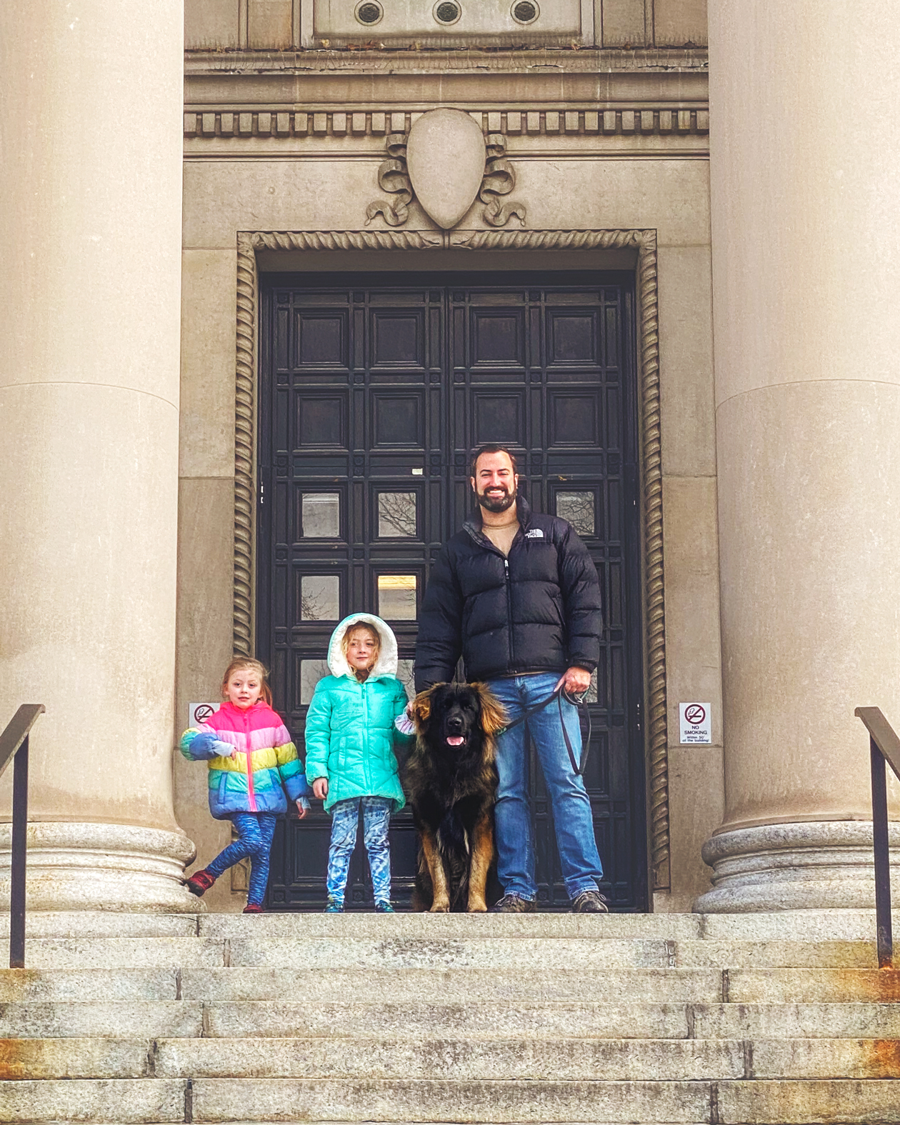 A man and two young girls stand with a dog on the steps of a large building