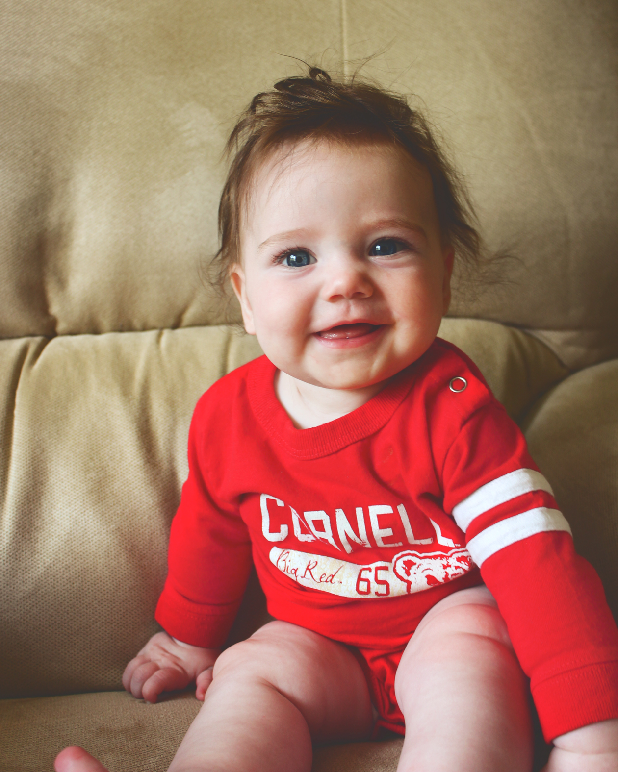 A baby girl wearing a red and white Cornell onesie sitting on a beige couch