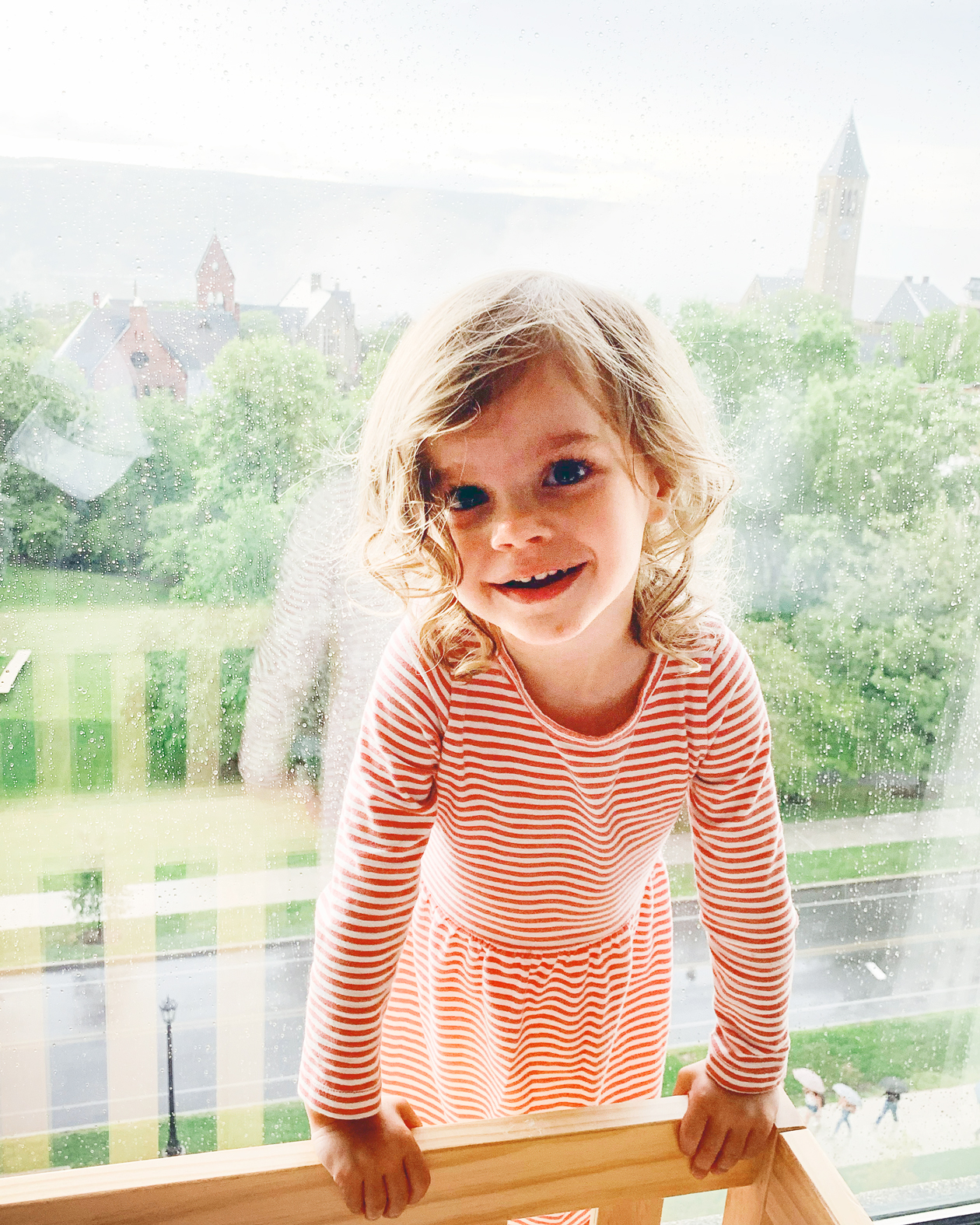A toddler girl wearing a red and white striped dress standing in front of a glass window with trees and buildings behind her