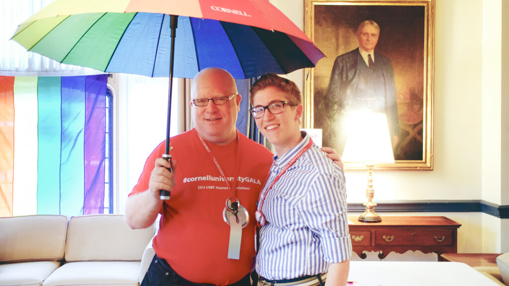 Two Cornell alumni stand under a rainbow umbrella at reunion.