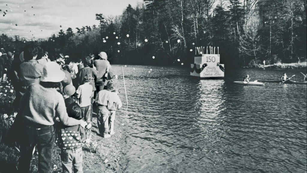 A B&W photo of the 150th birthday party for Beebe Lake, with a crowd on the shore and a cake-shaped float on the water
