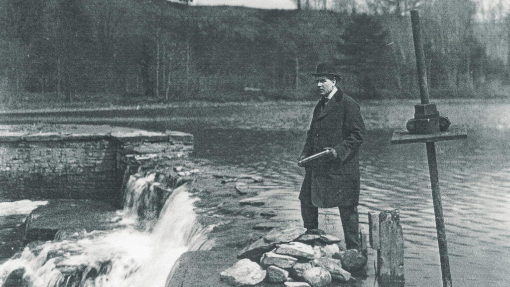 A man in an overcoat and bowler hat stands at the edge of Beebe Lake dam in a scene from the silent film "Mysteries of Myra"