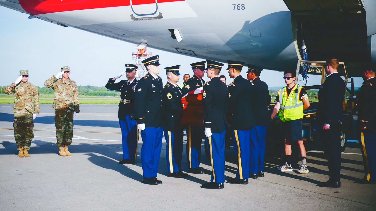 A military honor guard salutes the remains of a fallen service member on the runway of an airport next to an airplane.