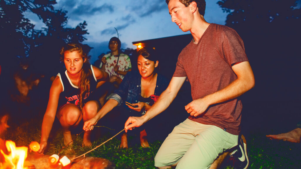 A group of college students roast marshmallows on a campfire in the evening.