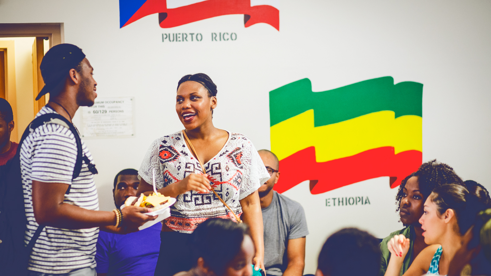 A group of people socialize in a room with the Puerto Rico and Ethiopia flags painted on the wall.