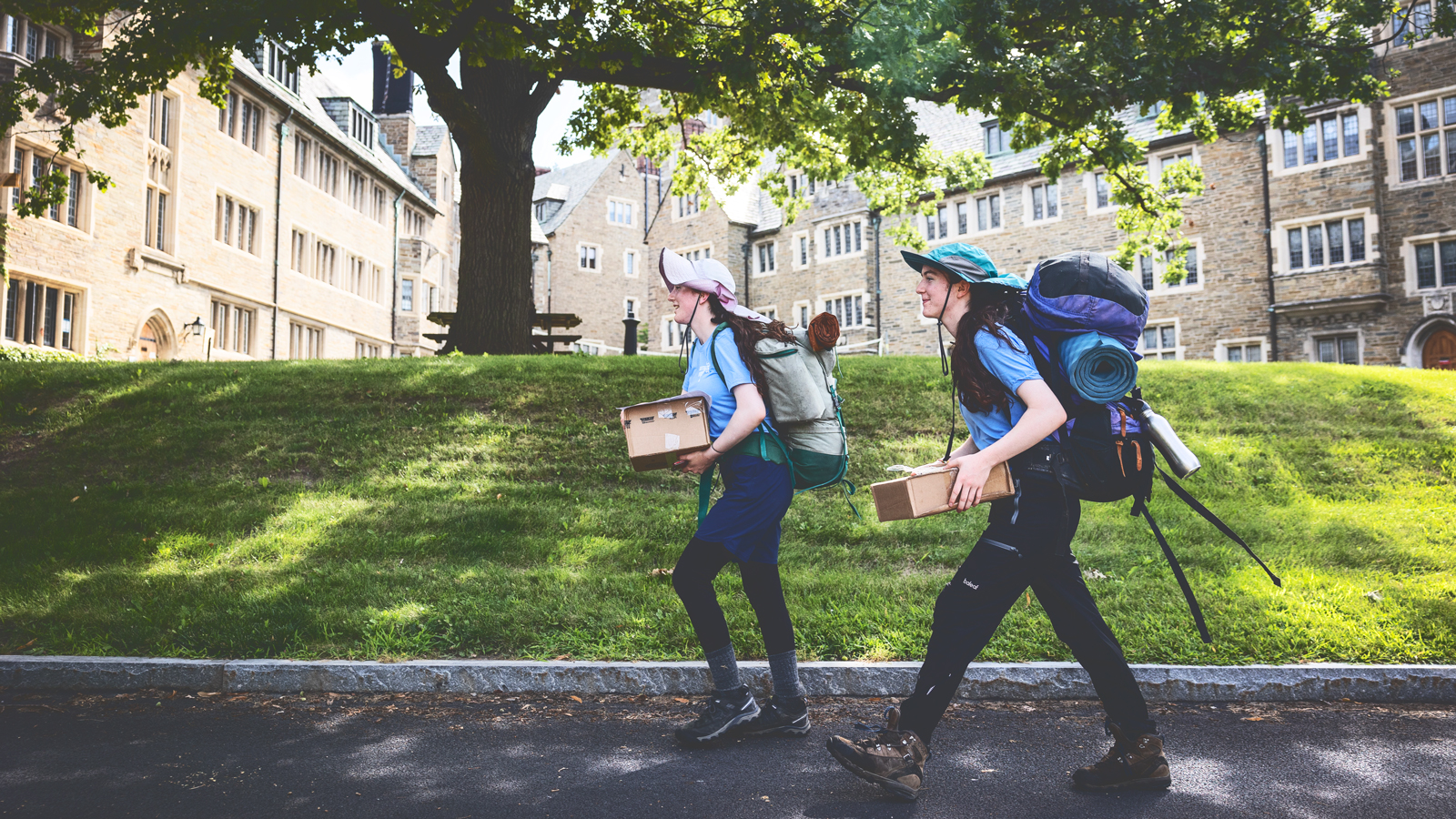 Two people wearing backpacks and holding boxes walk on the Cornell University campus.