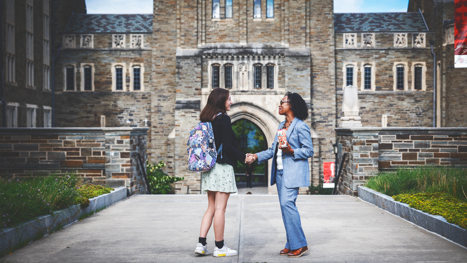 Two women shake hands on a sidewalk in front of a building on the Cornell University campus.