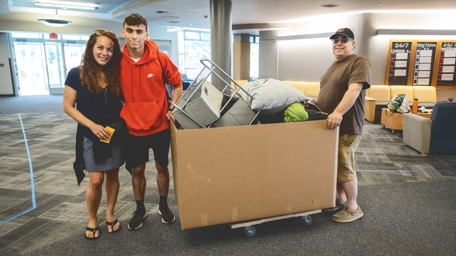A college student and his parents push a large box filed with furniture and pillows in a Cornell University dormitory.