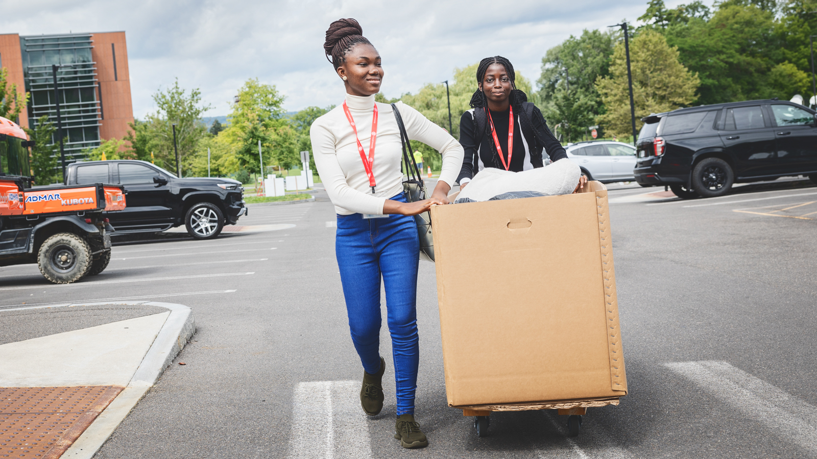 Two female Cornell students push a large box on wheels through a parking lot.