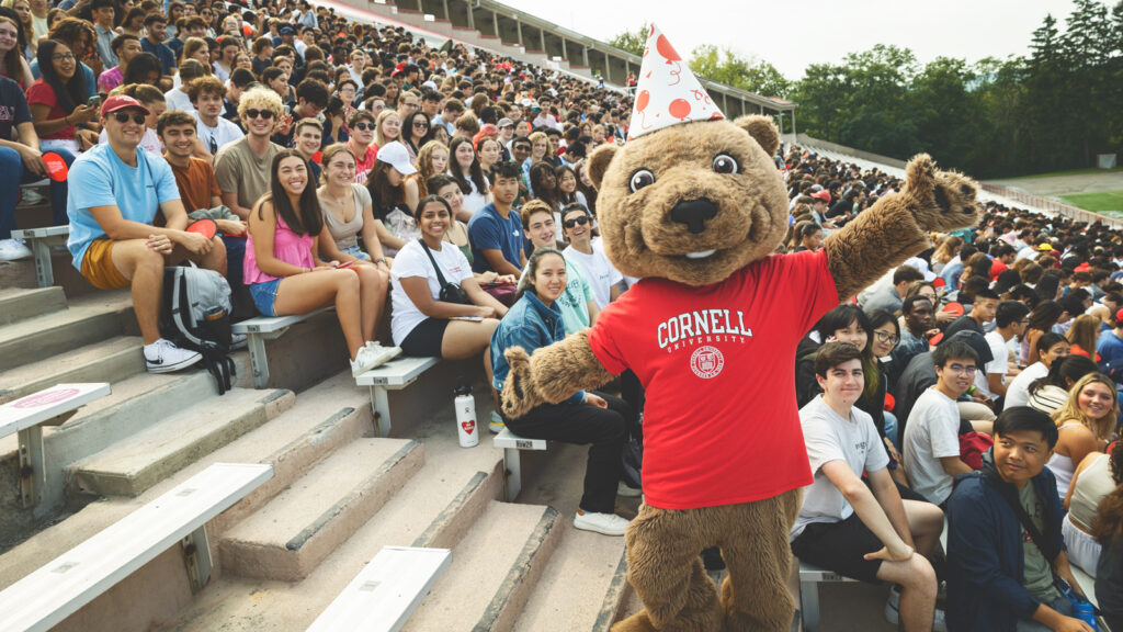A person in a bear costume stands with arms outstretched in front of a large crowd of students sitting in bleachers at a sports stadium at Cornell University.