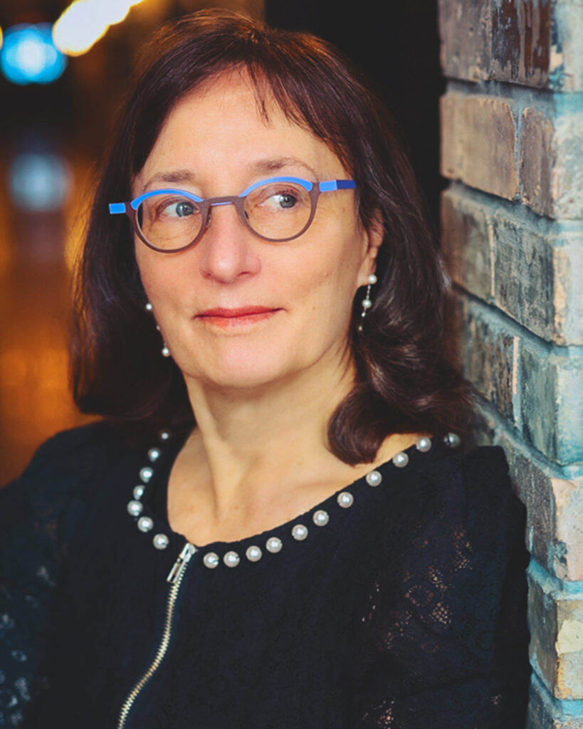 Author Julie Schumacher who has brown hair, blue-framed eyeglasses and is wearing a black sweater
