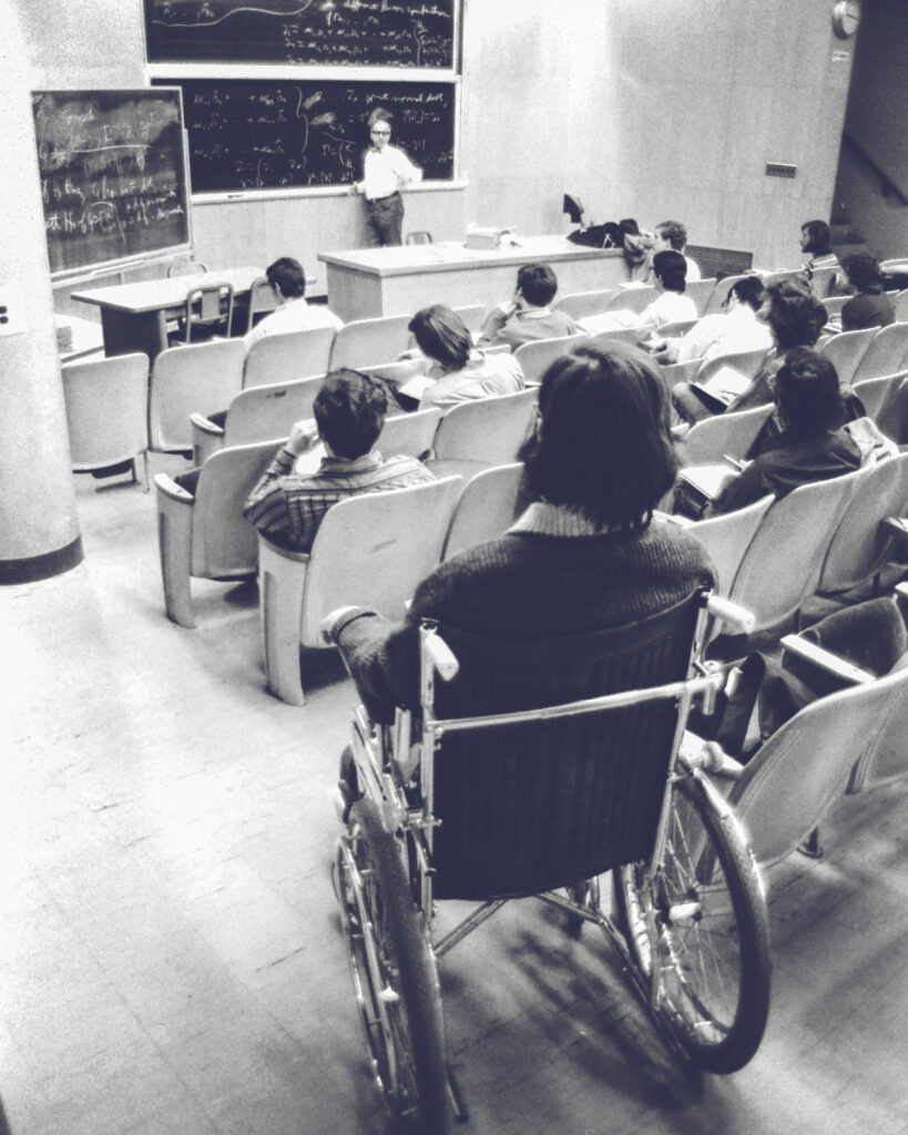 A man in a wheelchair attends a college lecture course surrounded by other students in seats.