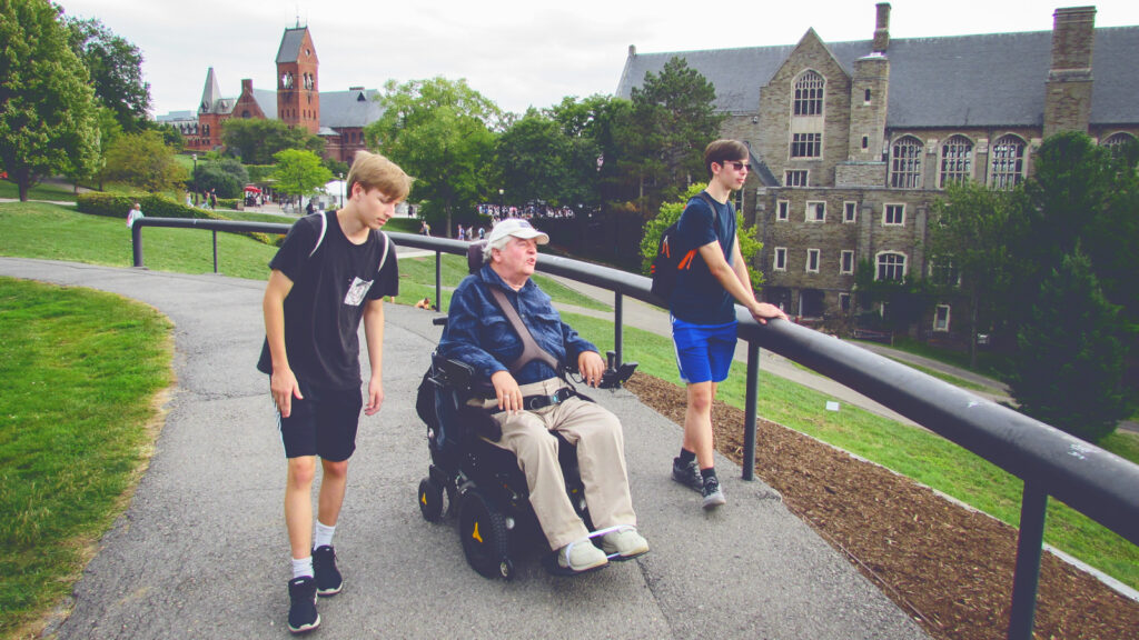 A man in a motorized wheelchair and two teenage boys tour a college campus.