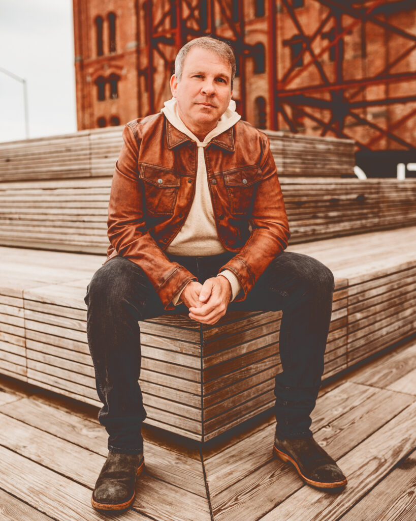 Steve Salm wearing a tan jacket and dark jeans sitting on a city rooftop