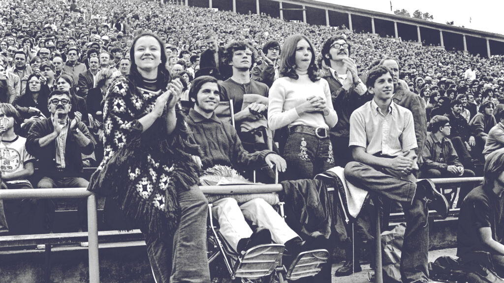 A paralyzed man in a wheelchair attends a college football game surrounded by other spectators.