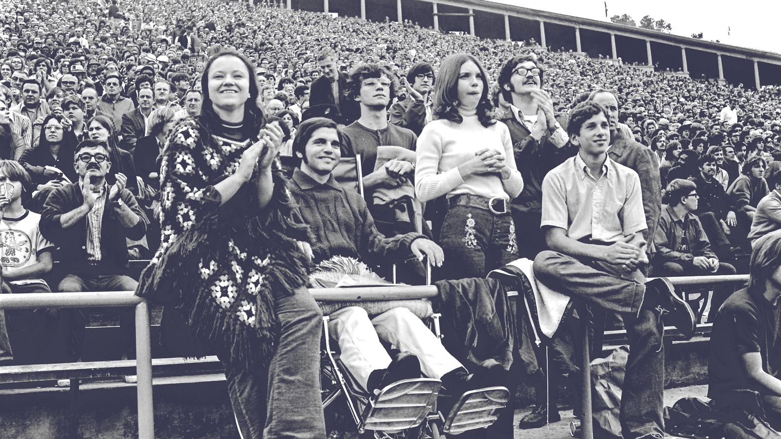 A paralyzed man in a wheelchair attends a college football game surrounded by other spectators.
