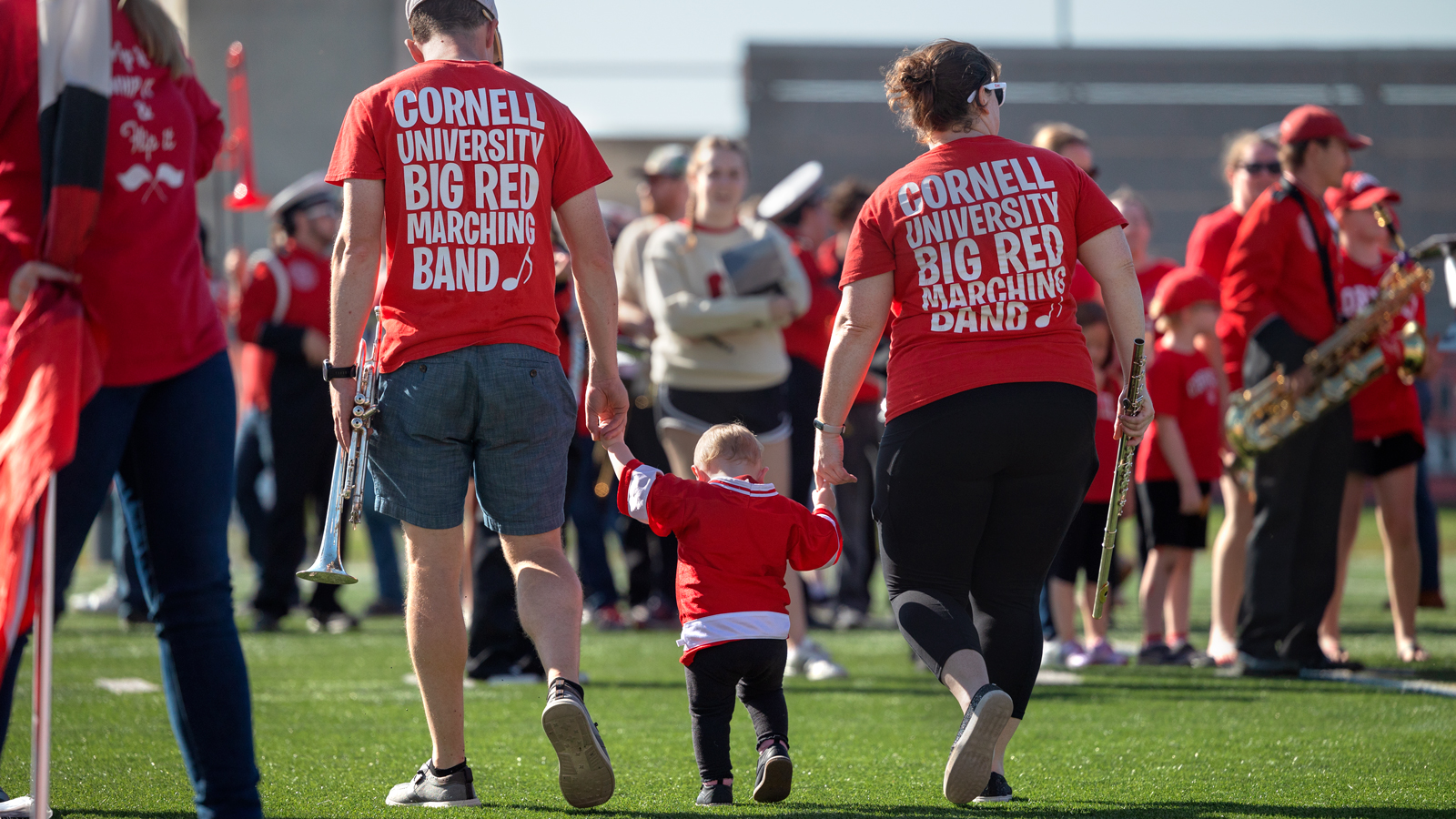 Two Big Red Marching Band alums walk on Schoellkopf Field with a small child