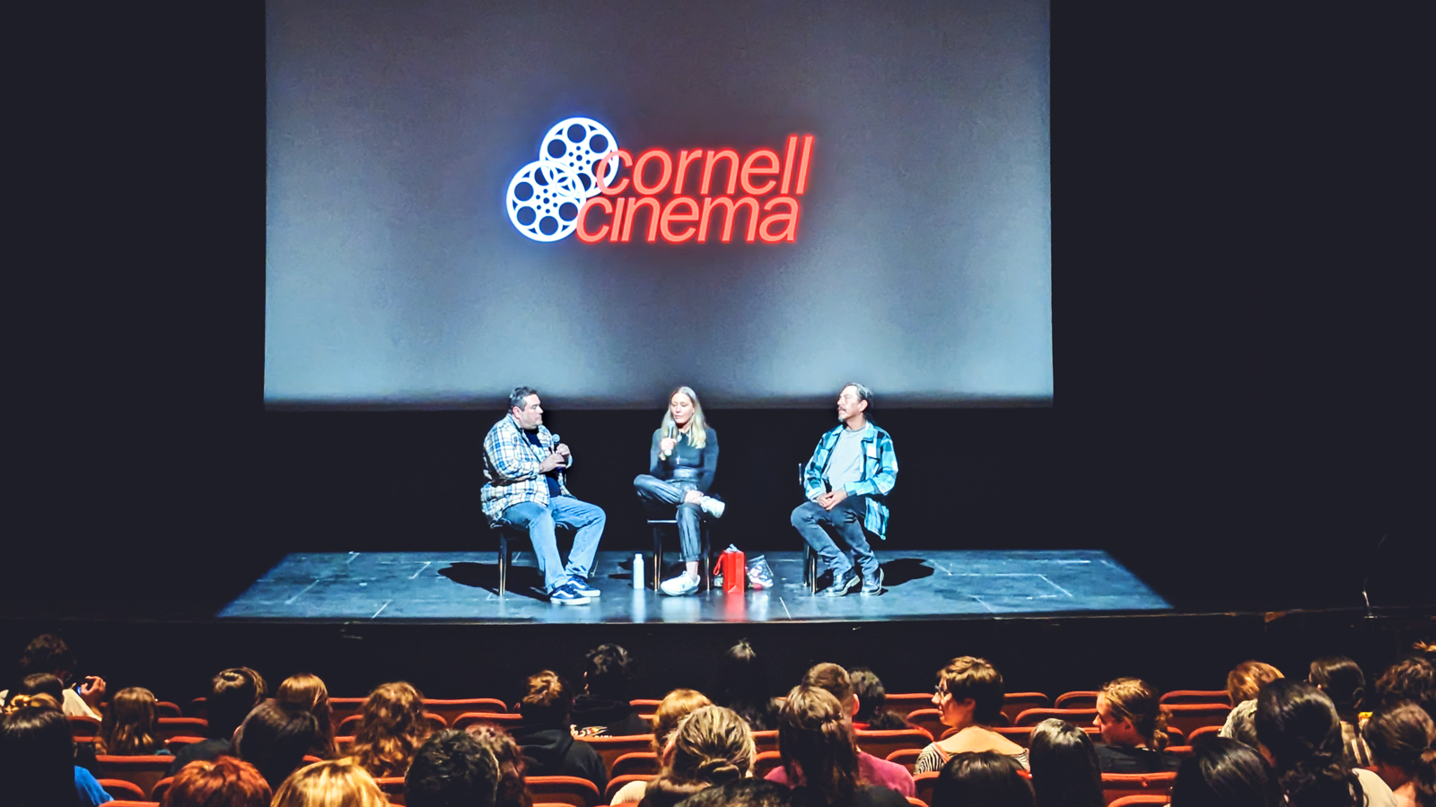 Three people in chairs on a stage talking to an audience with a movie screen behind them that says Cornell Cinema
