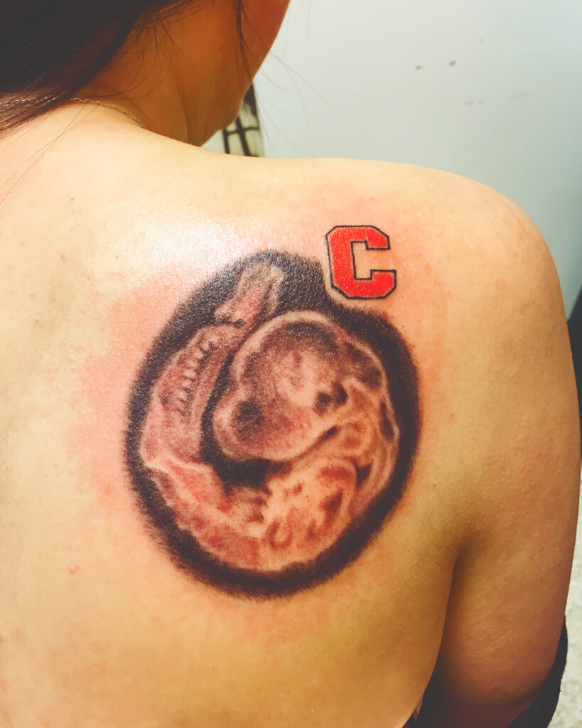 A tattoo of a red C and a pink mutant mouse embryo on a woman's right shoulder.
