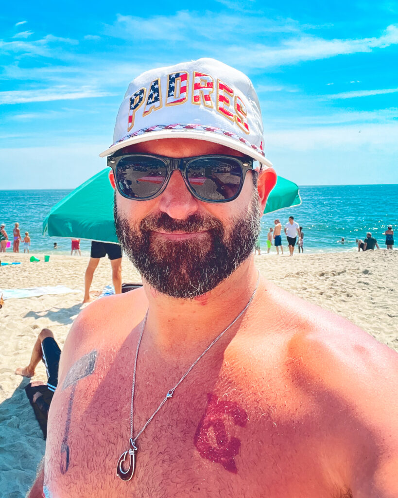 A man standing on a beach showing a Cornell University tattoo on his chest.