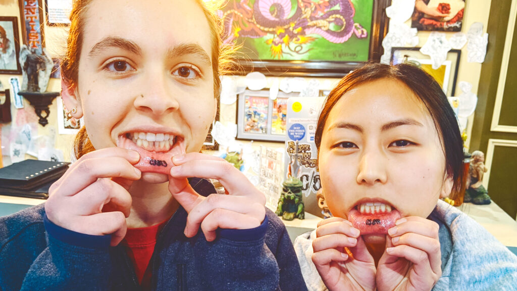 Two young women showing tattoos of numbers and letters on the inside of their lower lips
