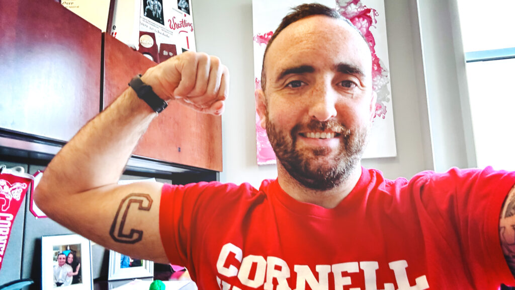 Cornell wrestling coach Mike Grey shows a C tattooed on his arm while wearing a red Cornell Wrestling t-shirt
