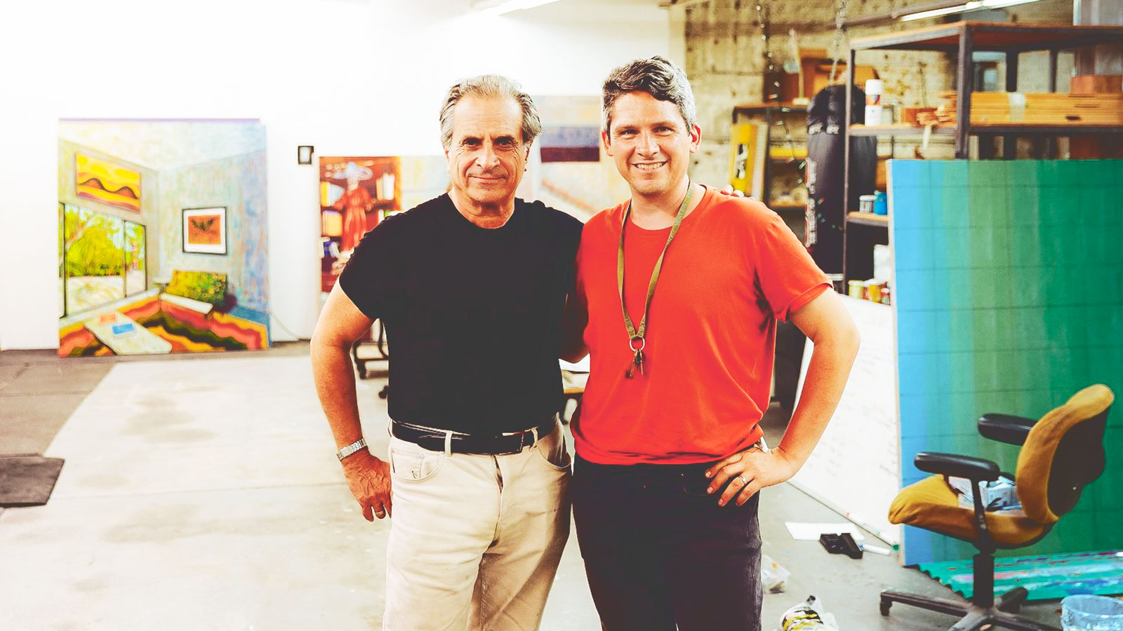 Gordon Sander and JJ Manford stand next to each other for a photo in a large art studio with several large colorful paintings in the background