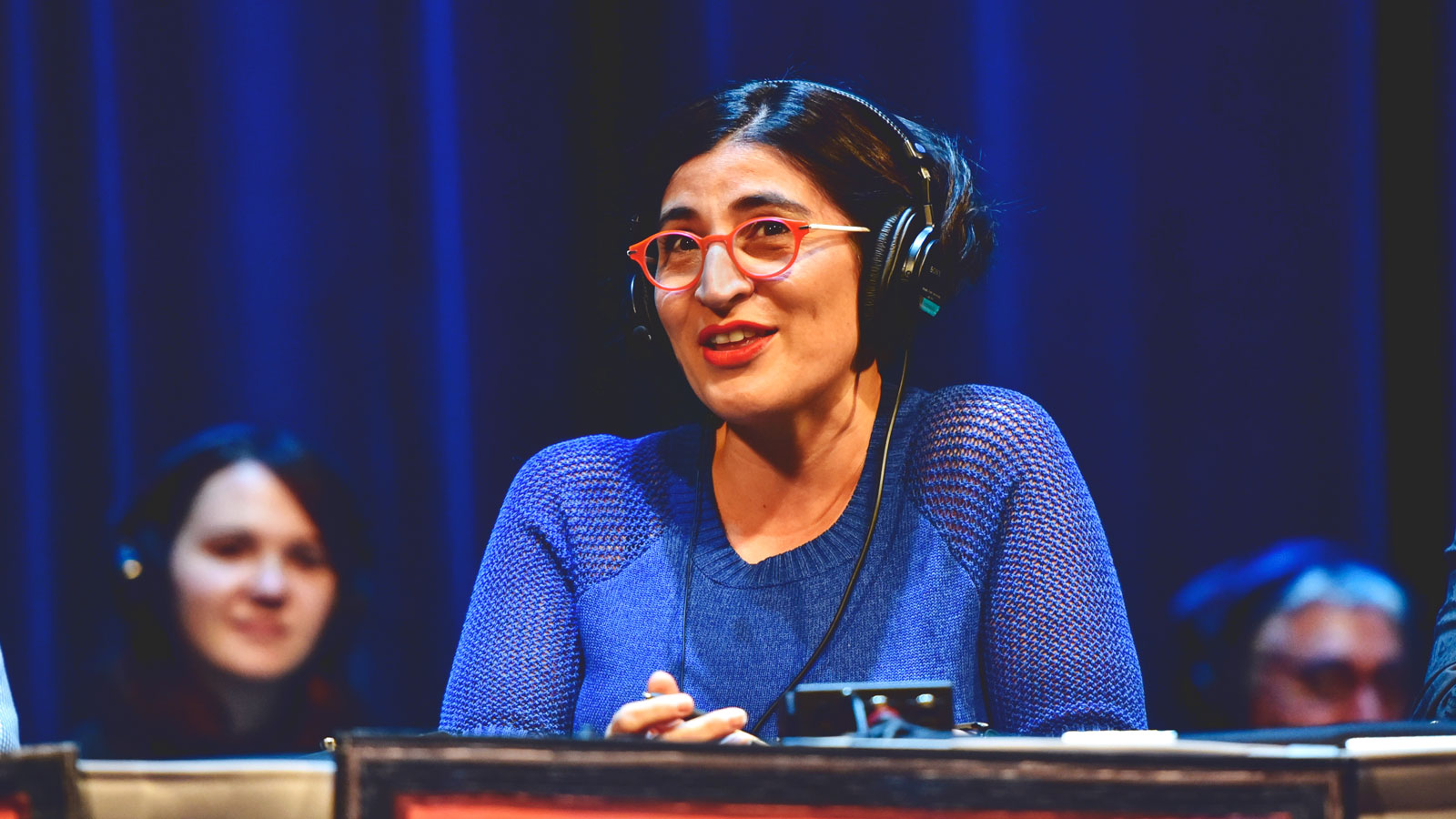 Comedian Negin Farsad wearing a blue dress, red glasses and a pair of headphones on a live radio program